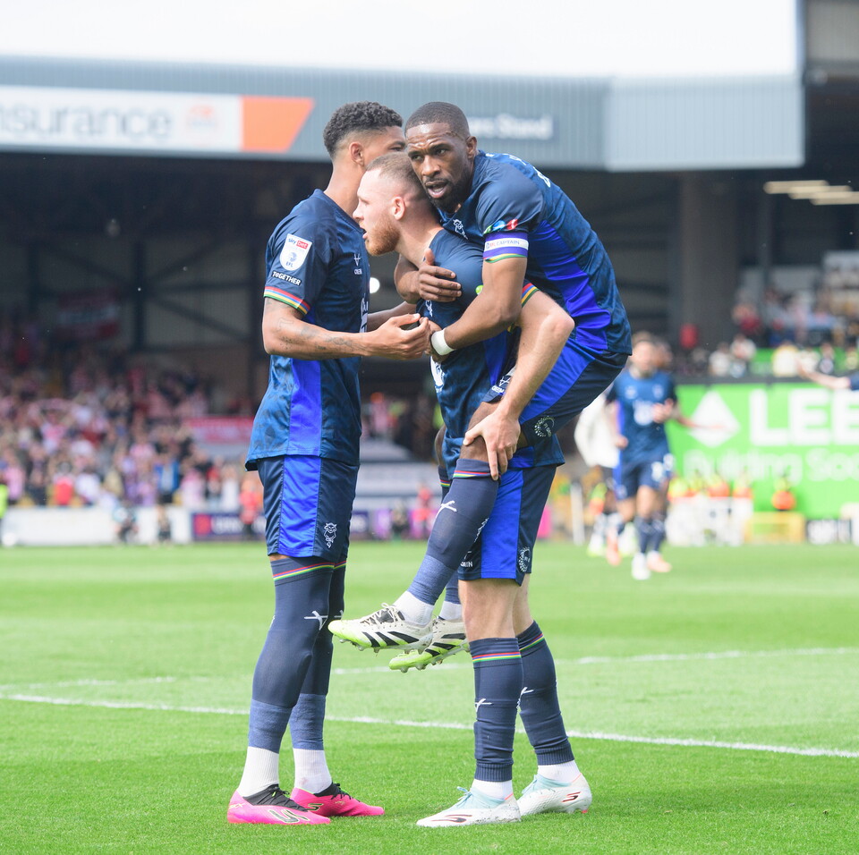 Three players in blue football kit celebrate a goal.