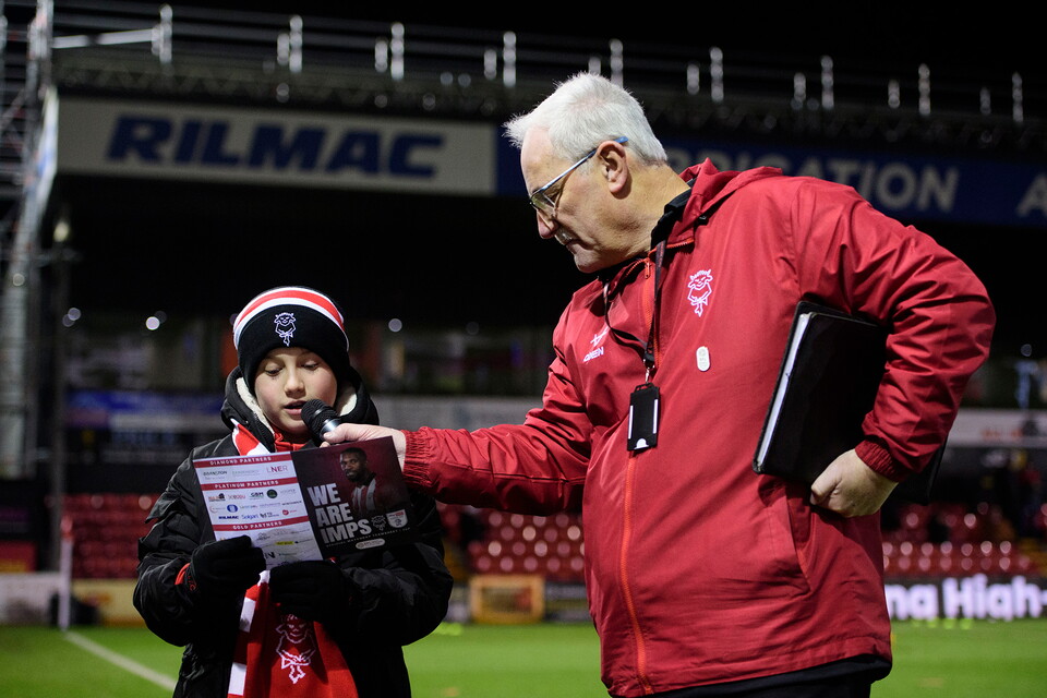 A child and an older man stand on a football field. The child reads from a card while the man, in a red jacket, holds a microphone and smiles gently.