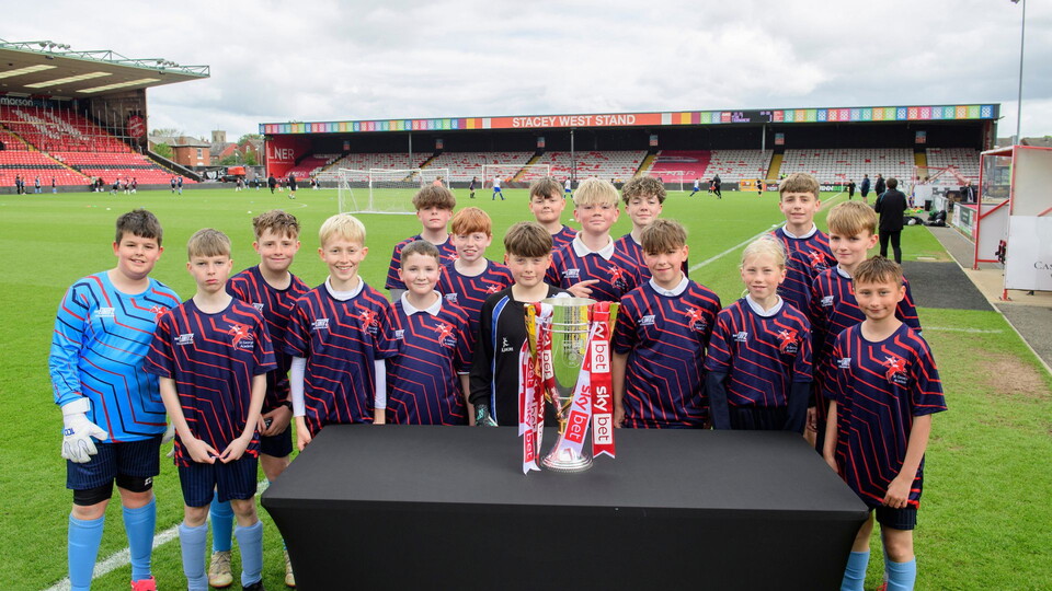 Young players from a team in blue and red kits pose proudly by a trophy on a football pitch, with a colorful stadium stand and cloudy sky in the background.
