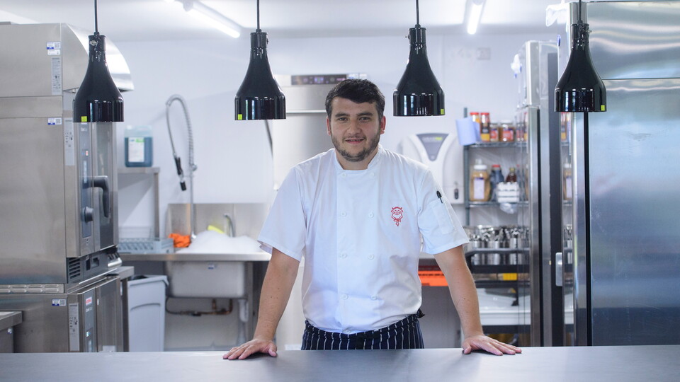 A chef stands at a counter, with a kitchen behind him.