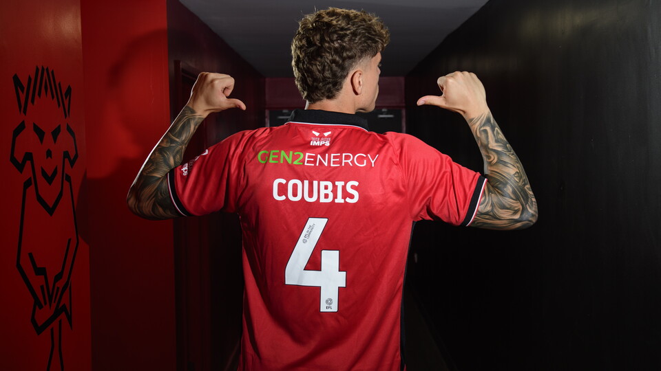 A man points to the back of his red shirt which reads "COUBIS 4". He is in a darkened room.