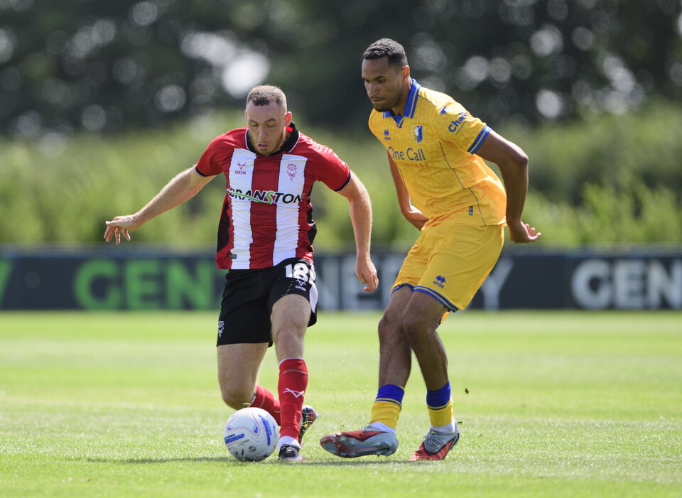 Ben House, wearing a red and white football top and black shorts, kicks a football