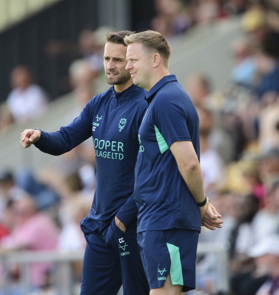 Two men in navy blue t-shirts stand on the side of a football pitch