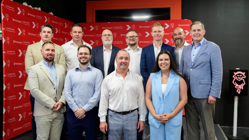 A group posing together in front of a red backdrop featuring a red Lincoln City crest. They are dressed in business casual attire and arranged in two rows.