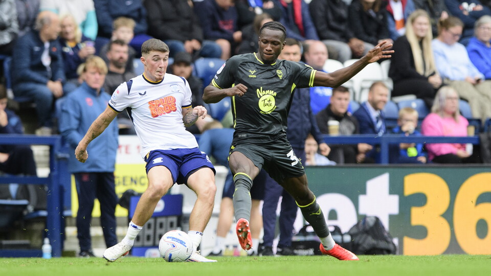 A man in a white football kit challenges for the ball with a man in a black football kit