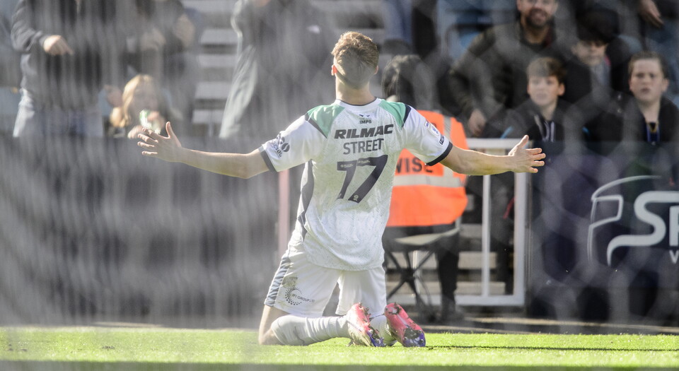 A man in a white football kit holds his arms in celebration after scoring a goal. He is facing away from the camera
