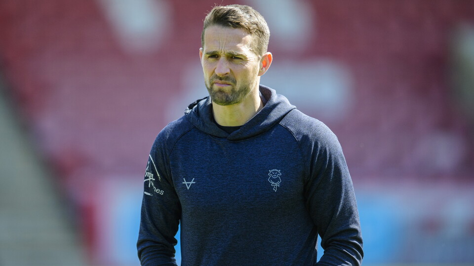 Chris Cohen stands on a football pitch before a game. He is looking left and wearing a blue hoody with the Oxen logo and the Lincoln City imp logos on.