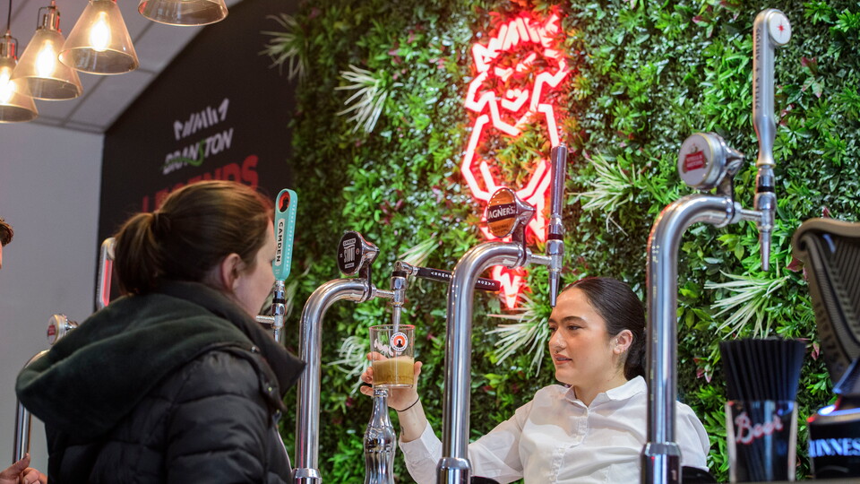 A view of the bar in the Legends Lounge. A woman wearing a white shirt pours a pint while another woman, wearing a black coat, waits.