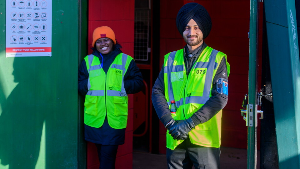 Two stewards, both wearing yellow high visibility jackets, stand at a turnstile entrance to the LNER Stadium. They are both smiling as they prepare to welcome supporters into the ground.
