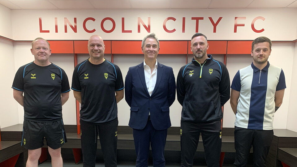 Marc Tracy, Jon Pepper, Jason Futers, Calum Oakenfold and Matt Murgett post in the changing room at the LNER Stadium. Above them is a sign which reads Lincoln City FC