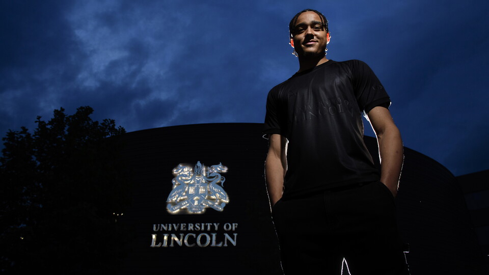 Jovon Makama wears Lincoln City's blackout third kit. He is stood in front of a building with the University of Lincoln logo on the wall.