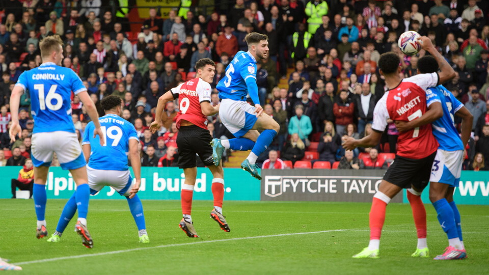 Bailey Cadamarteri heads a goal for Lincoln City. He is mid air, with a Stockport player alongside him, with the ball to his right heading towards the net.