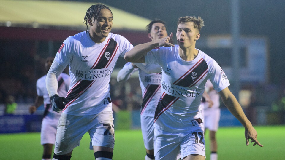 Jovon Makama and Erik Ring celebrate the latter's goal. Both are wearing white shirts with a black slash and running towards the camera.