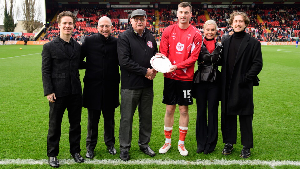 A group of people stand beside a football pitch. Stood in the middle is a man who is being presented with a silver salver.