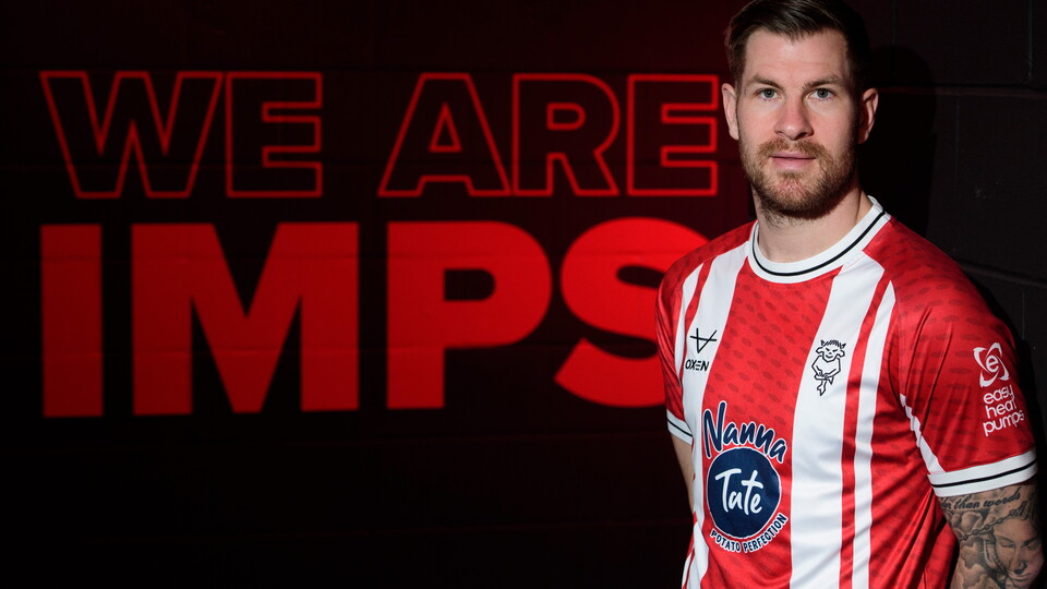 James Collins stands wearing a red and white Lincoln City top. In the background is a wall with the words We Are Imps on it.