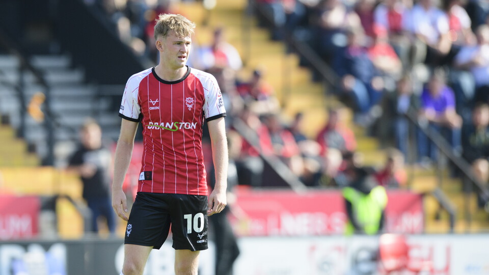 JJ McKiernan stands on a football pitch looking from the left of the image to the right. He is wearing a red football top with "Branston" on the front. In the background is a stand with people watching the match.