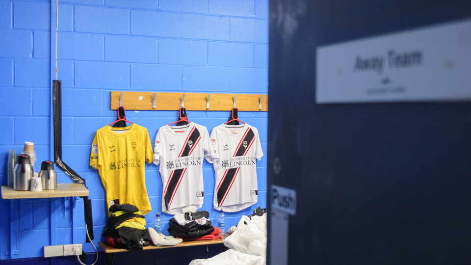 A view of three shirts hung up in a changing room, in the foreground is a door with a sign which reads "away team".