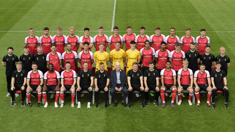 Lincoln City players and coaching staff pose for a team photo. Everyone on the front row is holding up a red sign which reads "Show racism the red card". There are three rows of people, with outfield players in red home shirts, goalkeepers in yellow shirts and staff in black kit.