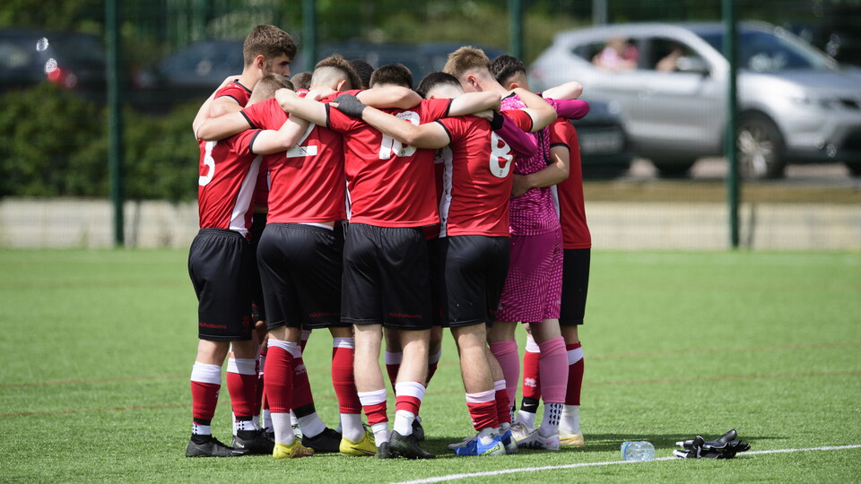 A group of boys form a huddle before playing football. They are wearing red shirts and black shorts.