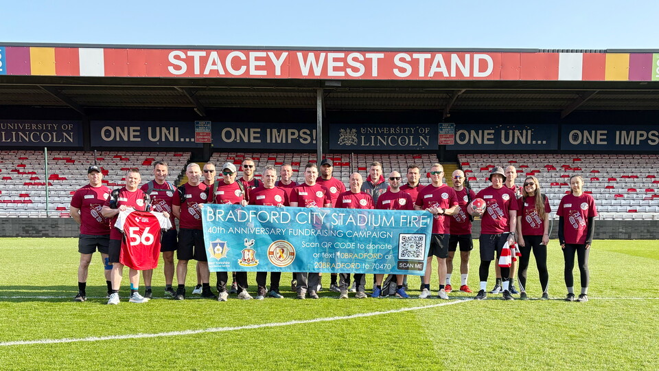 A group of people stand on a football pitch in front of a stand. On the roof of the stadium behind them is a sign which reads "STACEY WEST STAND"