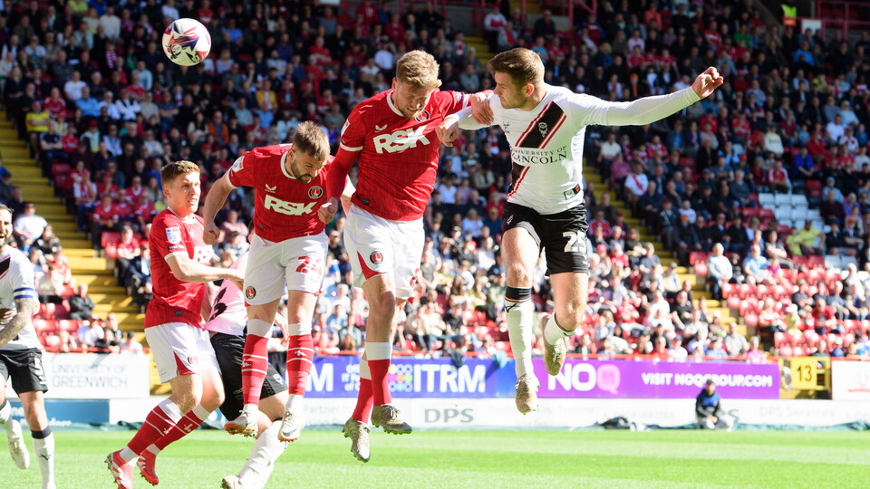 Four people are in the air as they challenge for the ball. The man on the right, wearing a white kit, has headed the ball towards goal.