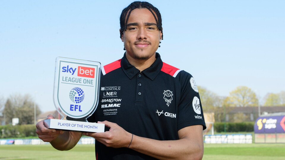Jovon Makama holds up a trophy which reads "Player of the Month". He is wearing a black polo shirt and stood against a blue sky. Behind him is grass and a football pitch.