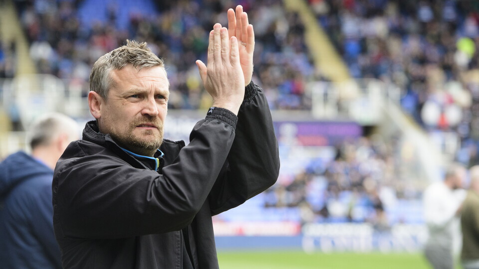 Michael Skubala holds his hands up to clap Lincoln City supporters. He is wearing a black rain jacket.