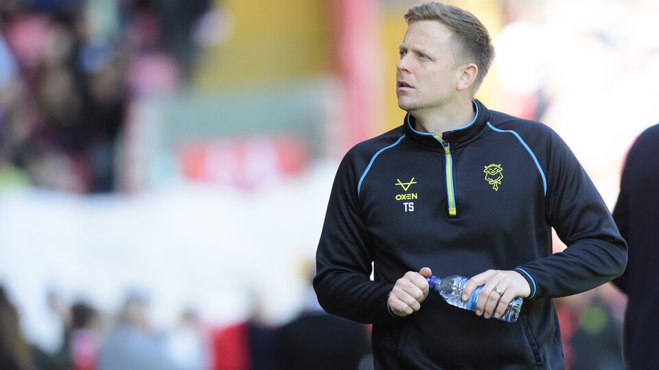 A man wearing all black tracksuit walks along the side of a football pitch. He's looking to his right.