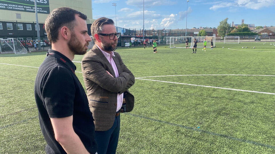 Two men watching a game of football on a football pitch. 