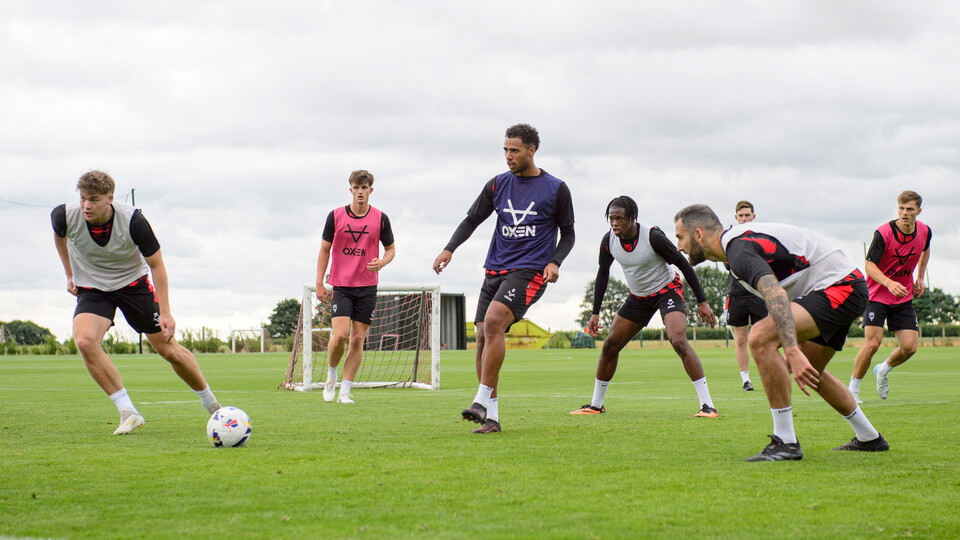 A group of men take part in football training on a grass pitch. The sky in the background is cloudy.