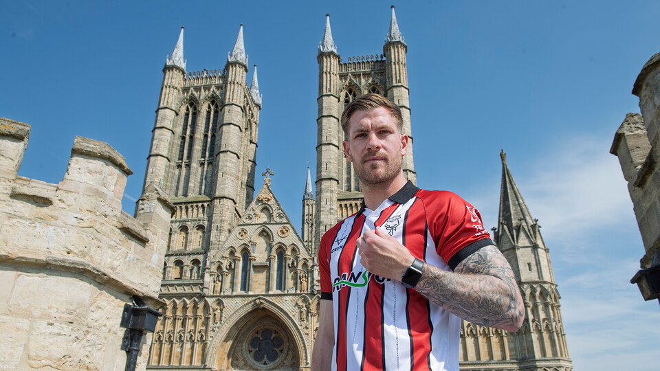 James Collins wearing a Lincoln City home shirt with Lincoln Cathedral in the background.