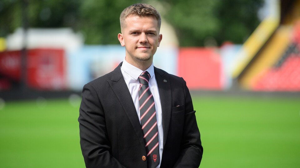A man stands in a white shirt, black and red tie, and black jacket.