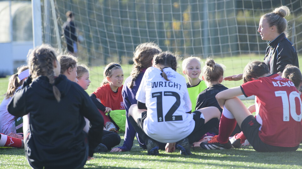 A young woman in a black tracksuit bends down to talk to a number of girls who are sat down. They are wearing a variety of football tops and taking a break from activities. It is a sunny day and in the background is a goal net.