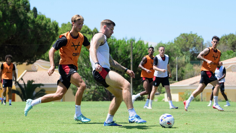 A football player wearing a white bib kicks a ball with his right foot as he is closed down by a player in an orange bib. There are players in the background.