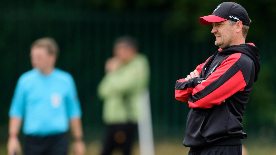 Lincoln City head coach watches a match from the sidelines. He is wearing a black and red hoody and baseball cap, and stood with his arms crossed. There are two people out of focus in the background.
