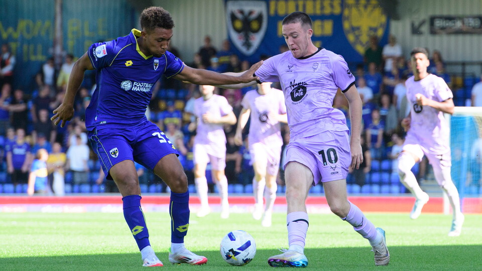 Jack Moylan, wearing an all violet shirt, has the ball at his feet as he dribbles with it at a man wearing a blue kit.