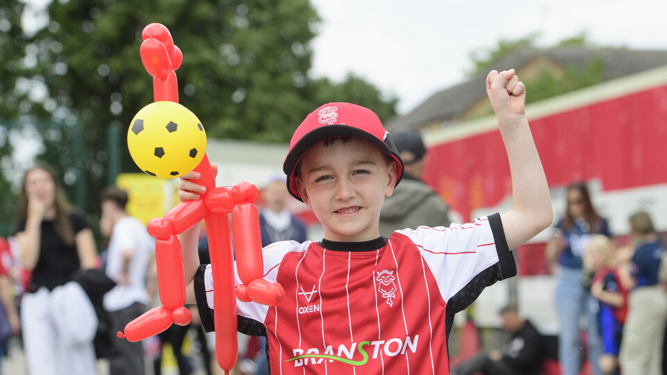 A child wears a replica Lincoln City shirt, he holds up a balloon animal.