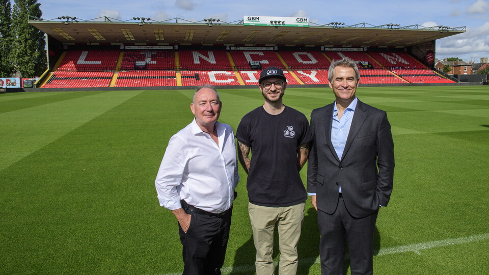 Three men stand in front of a football pitch with an empty stand in the background