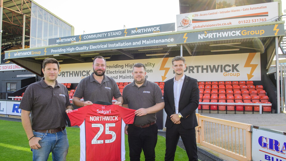 Four people stand holding a red shirt with Northwick 25 on the back