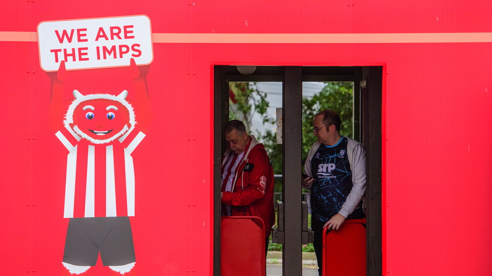 Two supporters enter the LNER Stadium through a turnstile. On the left is a mural featuring Poacher the Imp