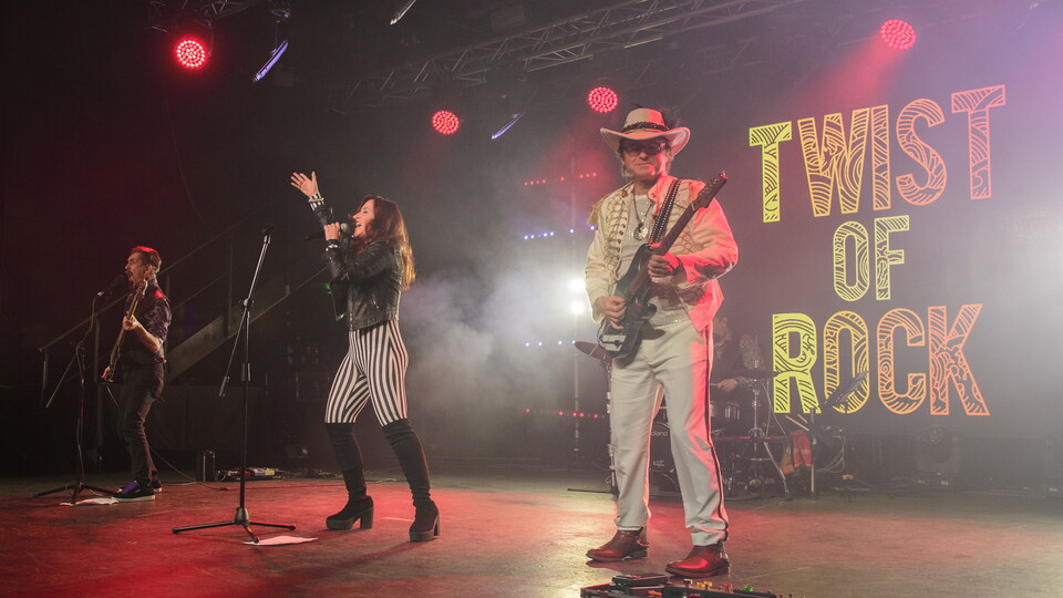 A band perform on a stage in front of a sign which reads "Twist Of Rock"