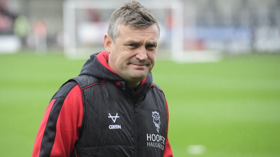 Michael Skubala wers a red hoody with a black gillet over it. He's stood in front of a football pitch.