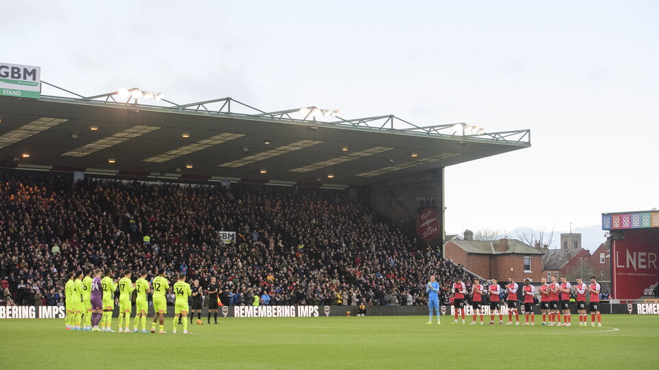 Lincoln City players line up for.a minute's applause. In the background are fans in a large stand as part of the LNER Stadium.