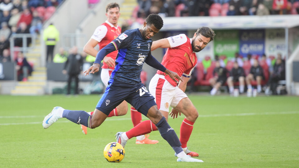 Justin Obikwu, wearing blue t-shirt and shorts, controls the football under pressure from a Rotherham United defender