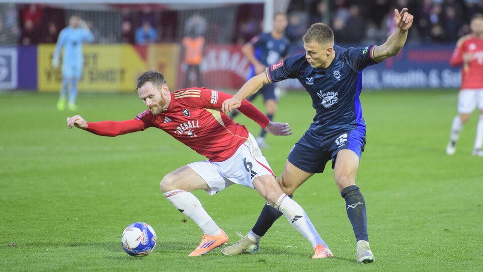 Lincoln's Erik Ring - wearing an all-blue football kit - challenges for the ball with a player in red shirt and white shorts.