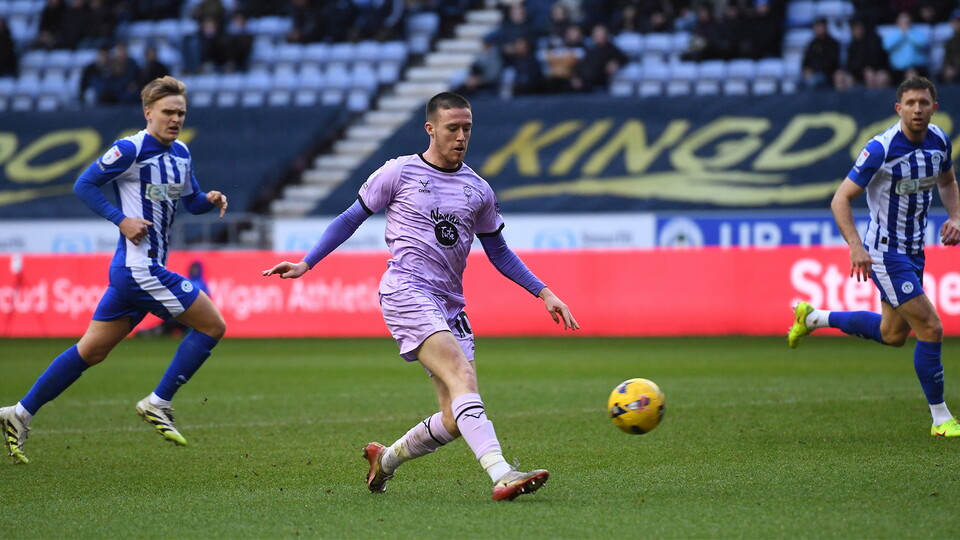 A player in a lilac coloured shirt clips a yellow football with two players in blue and white stripes watching on.