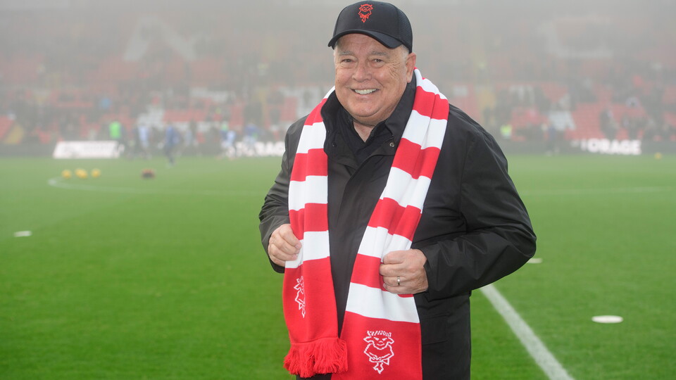 Ron Fowler stands wearing a black baseball cap. There is a red and white scarf around his neck.