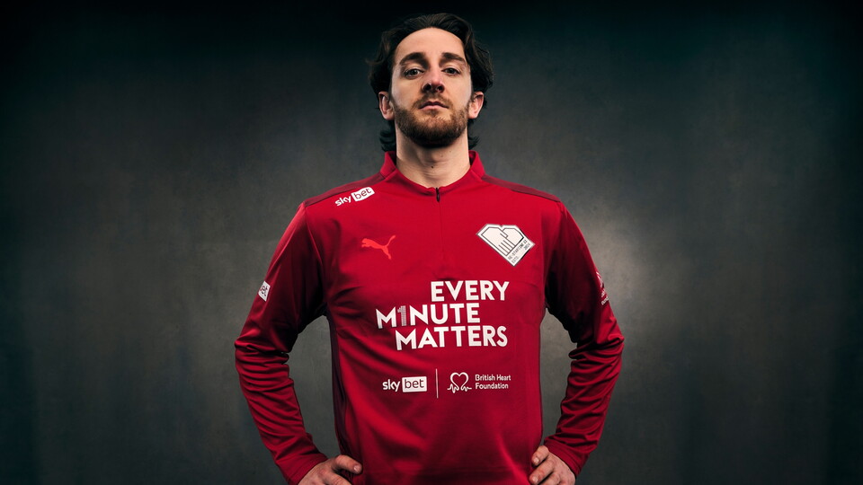 A man stands with hands on hips, wearing a red Puma jersey displaying "Every Minute Matters" and logos for Sky Bet and the British Heart Foundation.