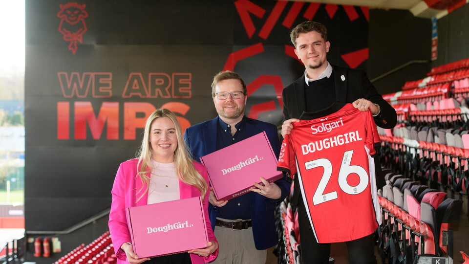 Three people stand in a stadium holding a pink box and a football shirt. The stadium has red seats and a 'We Are Imps' sign in the background.