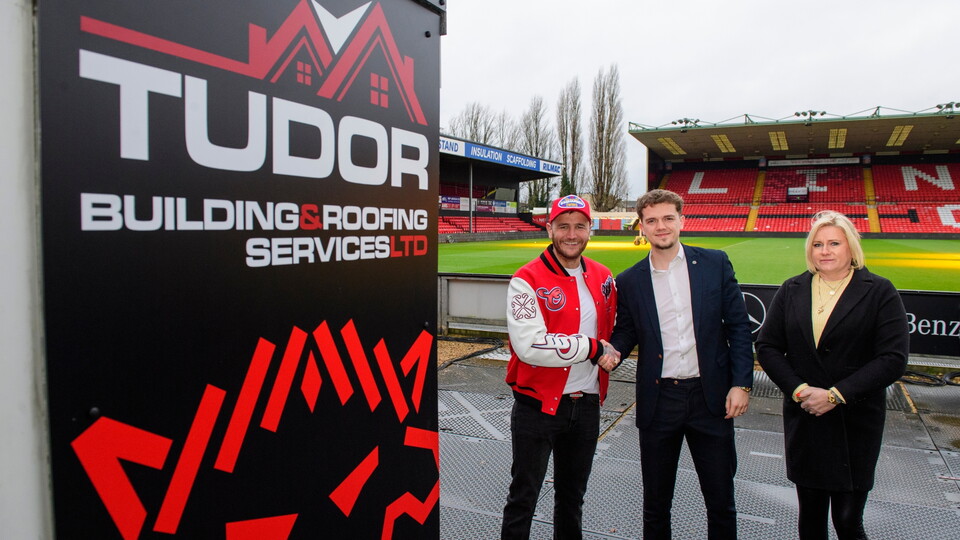 Three people stand smiling next to a Tudor Building & Roofing Services Ltd sign at a football stadium. The pitch and seating area are visible behind them.